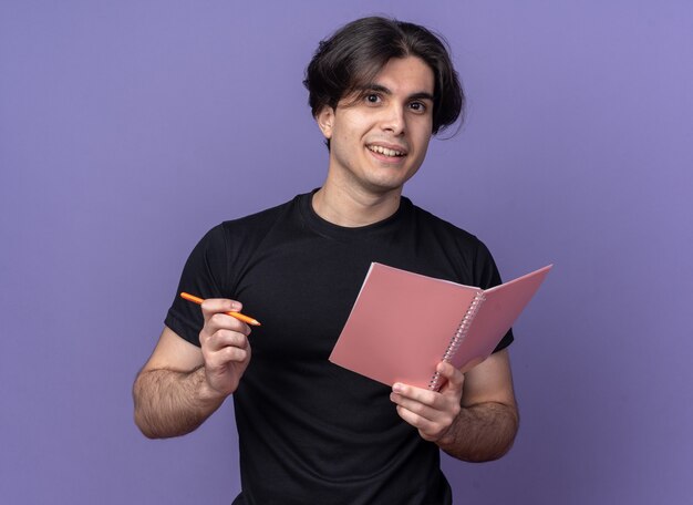 Souriant jeune beau mec portant un t-shirt noir tenant un cahier avec un stylo isolé sur un mur violet