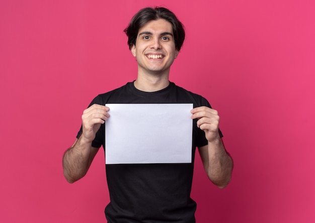 Souriant jeune beau mec portant un t-shirt noir tenant du papier isolé sur un mur rose