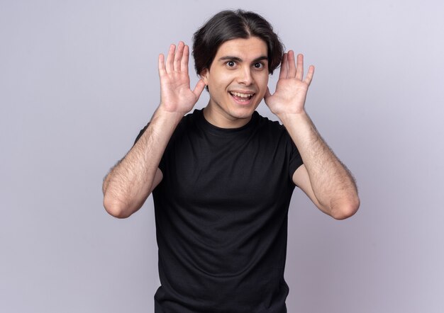 Souriant jeune beau mec portant un t-shirt noir tenant les mains autour des oreilles isolé sur mur blanc