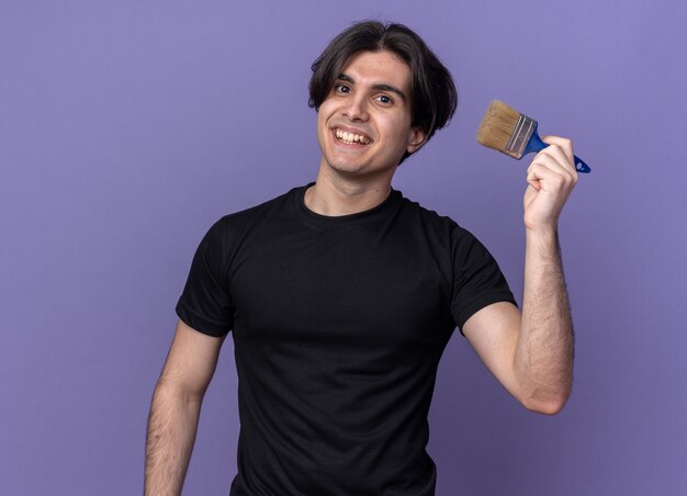 Souriant jeune beau mec portant un t-shirt noir tenant un pinceau isolé sur un mur violet