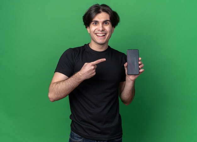 Souriant jeune beau mec portant un t-shirt noir tenant et points au téléphone isolé sur un mur vert