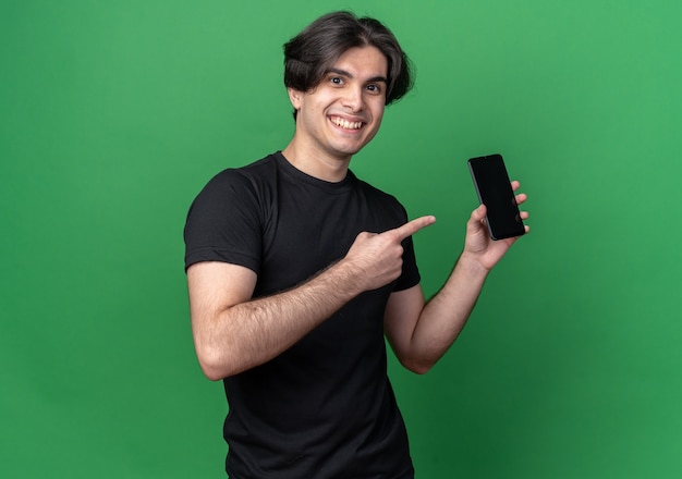 Souriant jeune beau mec portant un t-shirt noir tenant et points au téléphone isolé sur un mur vert