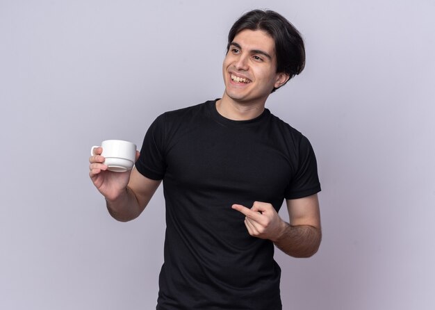 Souriant jeune beau mec portant un t-shirt noir tenant une tasse de points de café sur le côté isolé sur un mur blanc avec espace copie