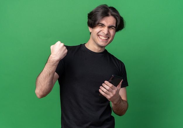 Souriant jeune beau mec portant un t-shirt noir tenant le téléphone et montrant oui geste isolé sur mur vert