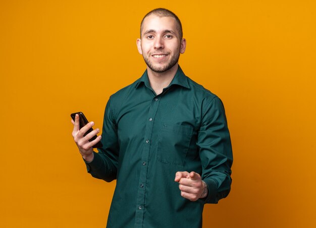 Souriant jeune beau mec vêtu d'une chemise verte tenant un téléphone et pointant vers l'avant