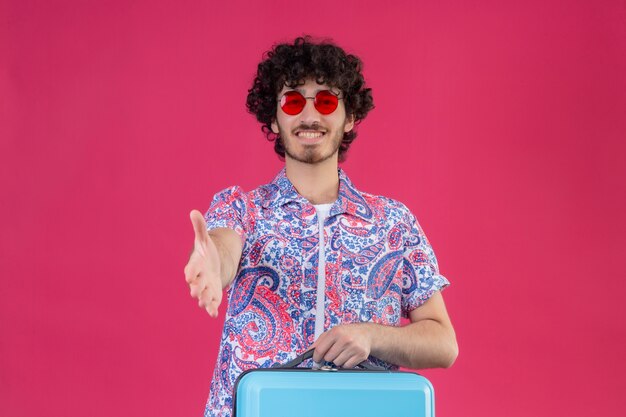 Souriant jeune beau voyageur bouclé homme portant des lunettes de soleil et étirant la main faisant des gestes salut sur mur rose isolé avec espace copie