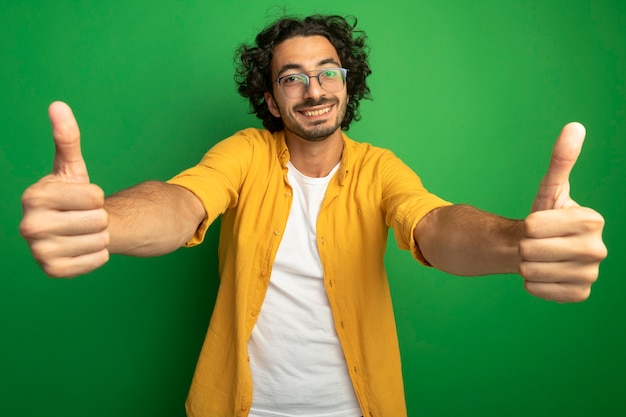 Photo gratuite souriant jeune bel homme caucasien portant des lunettes montrant les pouces vers le haut isolé sur le mur vert