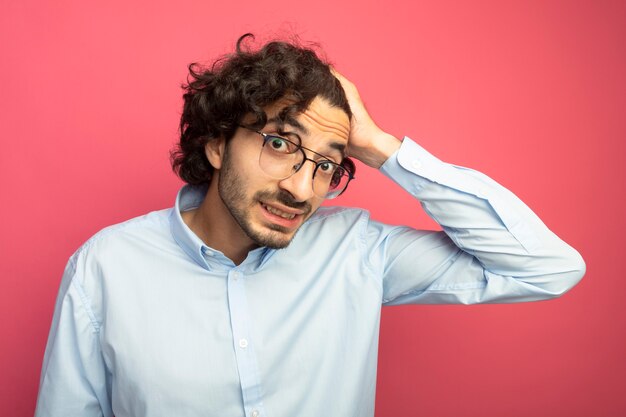 Souriant jeune bel homme caucasien portant des lunettes regardant la caméra en mettant la main sur la tête isolé sur fond cramoisi