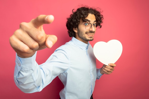 Photo gratuite souriant jeune bel homme caucasien portant des lunettes tenant la forme de coeur à la recherche et pointant la caméra isolée sur fond cramoisi