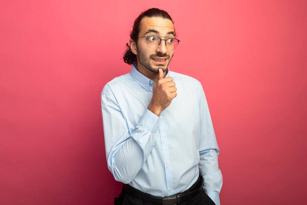 Souriant jeune bel homme caucasien portant des lunettes touchant le menton en gardant la main dans la poche à côté isolé sur fond cramoisi avec espace copie