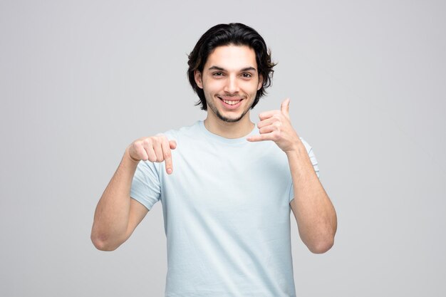 Souriant jeune bel homme montrant un geste d'appel pointant vers le bas tout en regardant la caméra isolée sur fond blanc