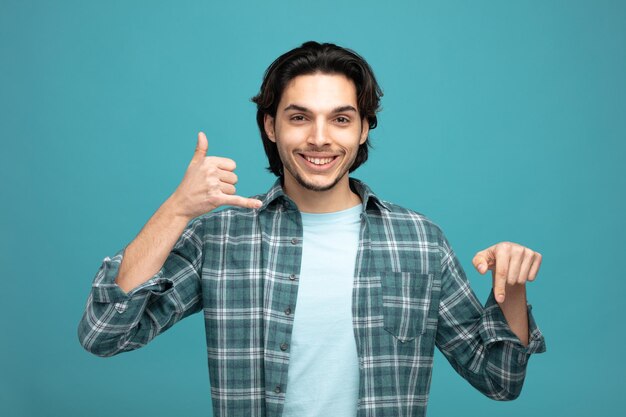 souriant jeune bel homme montrant un geste d'appel regardant la caméra pointant vers le bas isolé sur fond bleu