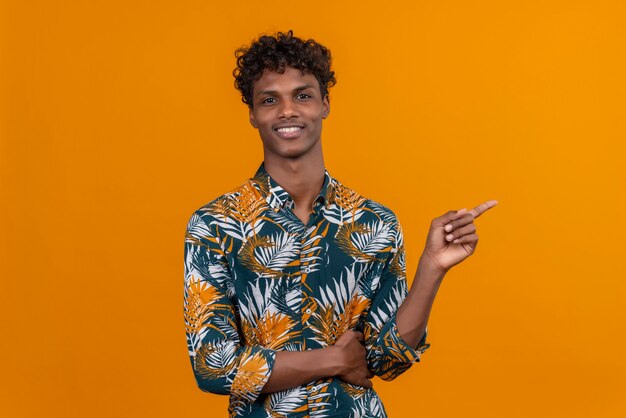 Souriant jeune bel homme à la peau foncée aux cheveux bouclés en chemise imprimée de feuilles montrant quelque chose avec l'index sur fond orange