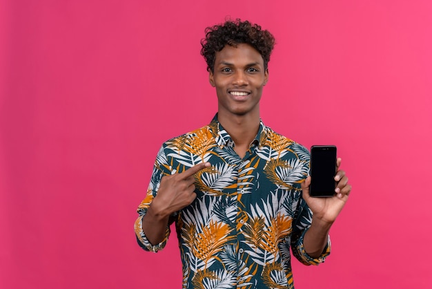 Souriant jeune bel homme à la peau foncée avec des cheveux bouclés en chemise imprimée de feuilles pointant sur le téléphone mobile avec l'index sur fond rose