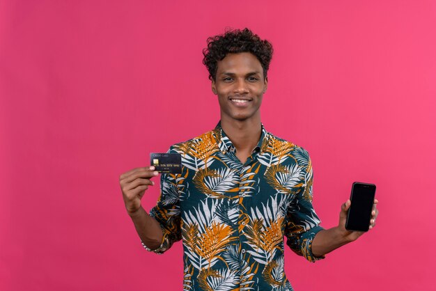 Souriant jeune bel homme à la peau sombre avec des cheveux bouclés en chemise imprimée de feuilles montrant un écran de téléphone intelligent vierge avec carte de crédit sur fond rose