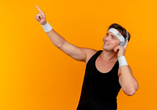 Souriant jeune bel homme sportif portant un bandeau et des bracelets avec un casque à la recherche et pointant vers le haut et en mettant le doigt sur des écouteurs isolés sur un mur orange