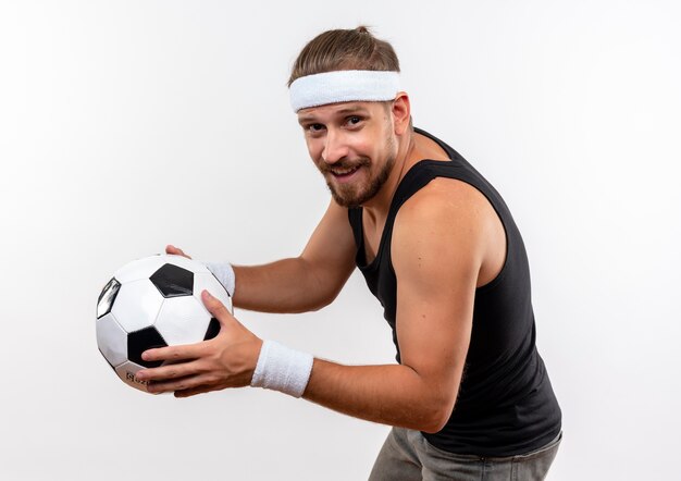 Souriant jeune bel homme sportif portant bandeau et bracelets debout en vue de profil tenant un ballon de soccer isolé sur un espace blanc