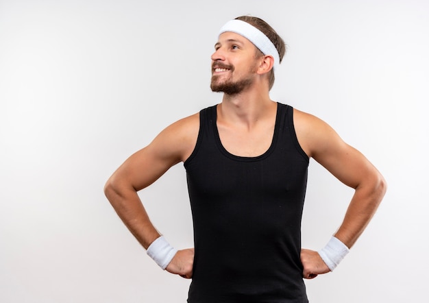 Souriant jeune bel homme sportif portant un bandeau et des bracelets en regardant le côté avec les mains sur la taille isolé sur un espace blanc