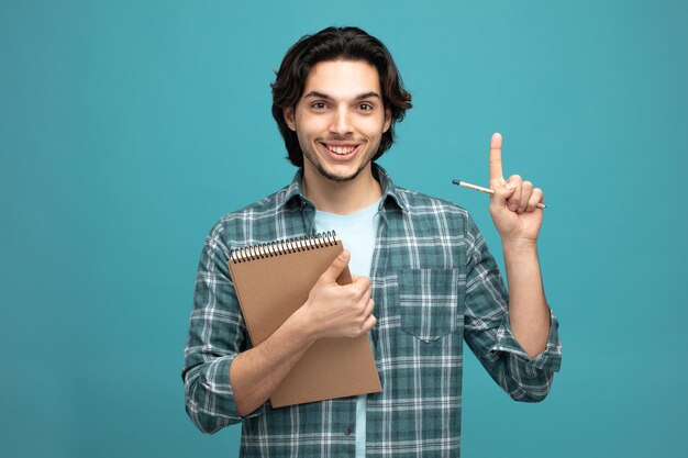 souriant jeune bel homme tenant un bloc-notes et un crayon regardant la caméra pointant vers le haut isolé sur fond bleu