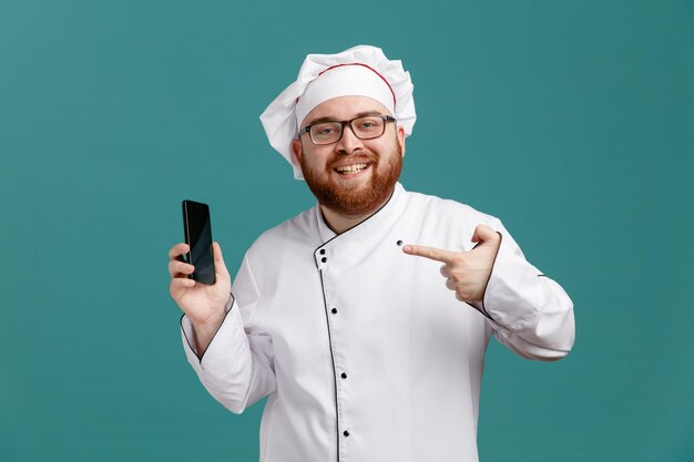 Souriant jeune chef masculin portant des lunettes uniformes et une casquette montrant un téléphone portable pointant dessus regardant la caméra isolée sur fond bleu