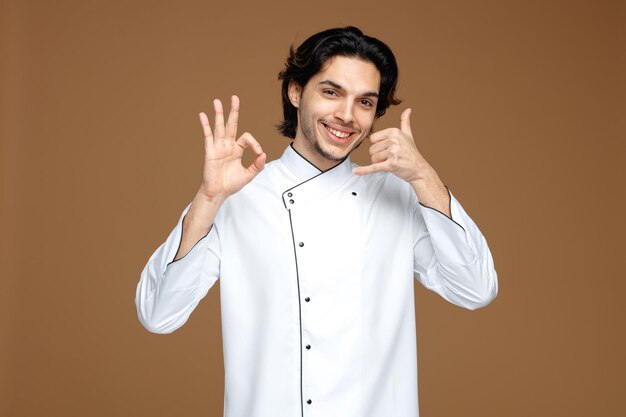 souriant jeune chef masculin portant l'uniforme regardant la caméra montrant le signe ok et le geste d'appel isolé sur fond marron