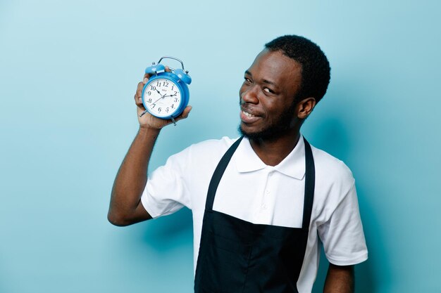 Souriant jeune coiffeur afro-américain en uniforme tenant un réveil isolé sur fond bleu