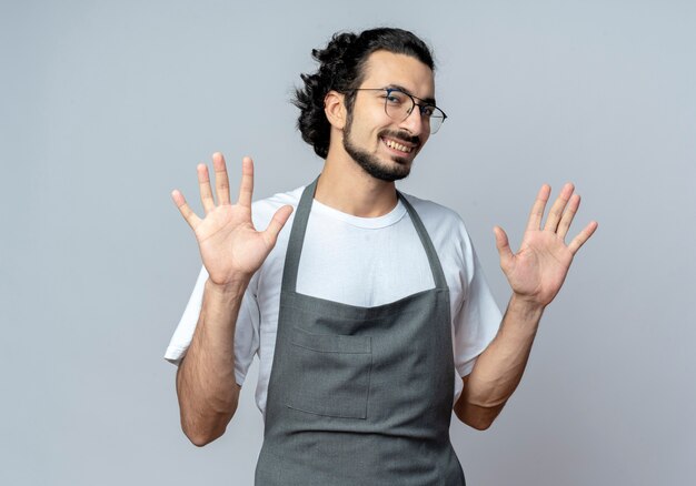 Souriant jeune coiffeur masculin de race blanche portant des lunettes et une bande de cheveux ondulés en uniforme montrant les mains vides sur fond blanc