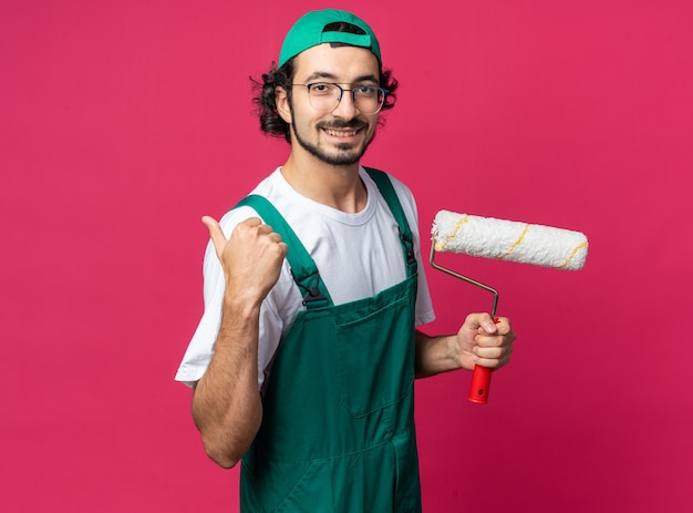 Souriant jeune constructeur homme portant un uniforme avec une casquette tenant une brosse à rouleau montrant le pouce vers le haut