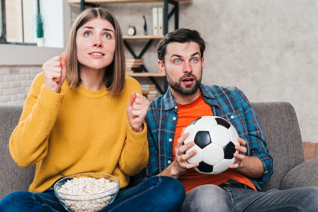 Photo gratuite souriant jeune couple assis sur un canapé en regardant le match de football