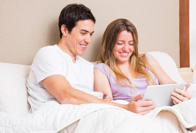 Souriant jeune couple assis sur le lit en regardant une tablette numérique