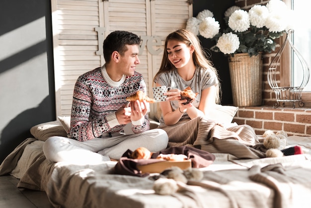 Souriant jeune couple assis sur le lit, tenant un croissant et un petit gâteau à la main
