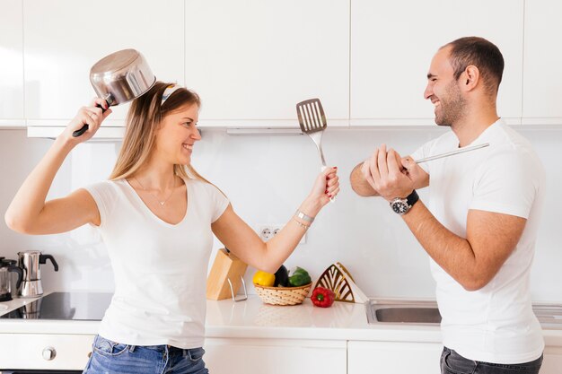 Souriant jeune couple se battre avec un ustensile et une spatule dans la cuisine