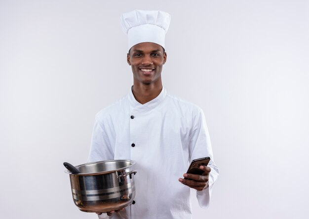 Souriant jeune cuisinier afro-américain en uniforme de chef détient casserole et téléphone sur fond blanc isolé avec copie espace