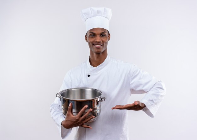 Souriant jeune cuisinier afro-américain en uniforme de chef tient une casserole et garde la main droite sur fond blanc isolé avec copie espace