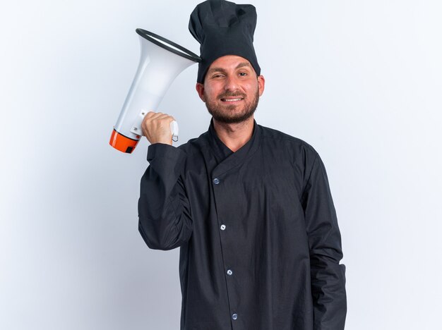 Souriant jeune cuisinier masculin de race blanche en uniforme de chef et casquette touchant la tête avec haut-parleur