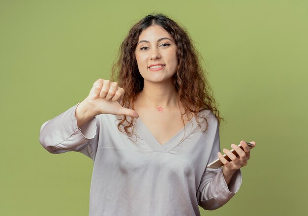 Souriant jeune employé de bureau jolie femme tenant le téléphone et son pouce vers le bas isolé sur vert olive