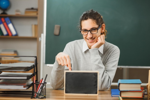 souriant jeune enseignant portant des lunettes assis au bureau tenant un mini tableau avec des outils scolaires en classe