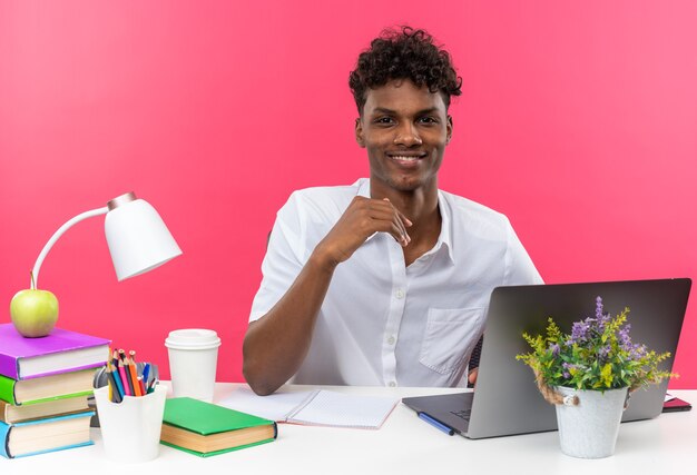 Souriant jeune étudiant afro-américain assis au bureau avec des outils scolaires à la recherche d'isolement sur le mur rose