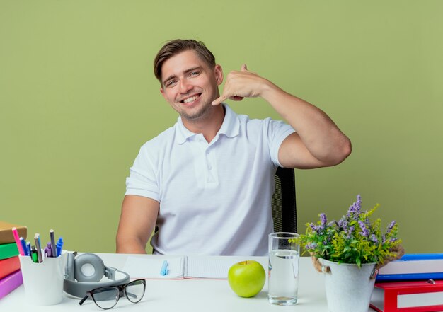 Souriant jeune étudiant beau mâle assis au bureau avec des outils scolaires montrant le geste d'appel téléphonique isolé sur vert olive