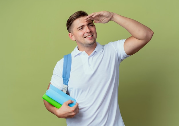 Souriant jeune étudiant beau mâle portant un sac à dos regardant à distance avec la main et tenant le livre isolé sur vert olive