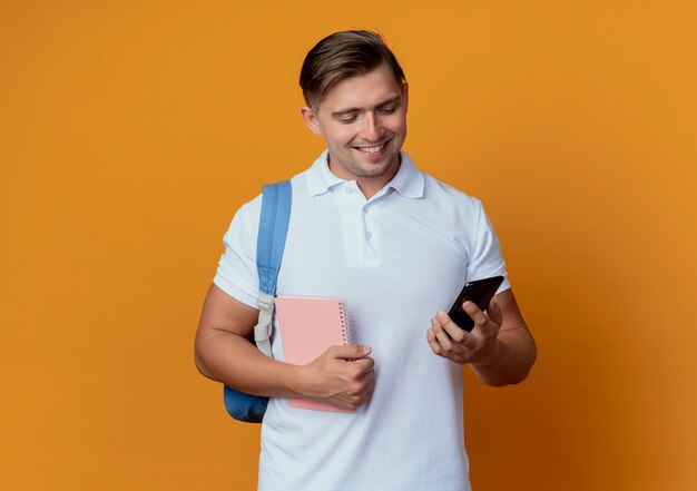 Souriant jeune étudiant beau mâle portant sac à dos tenant le cahier et regardant le téléphone dans sa main isolé sur orange