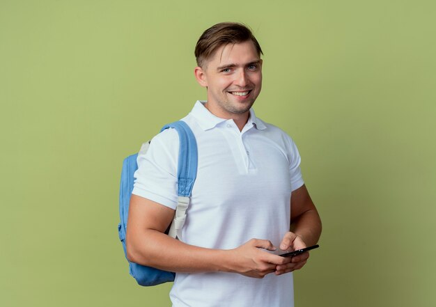 Souriant jeune étudiant beau mâle portant sac à dos tenant le téléphone isolé sur vert olive