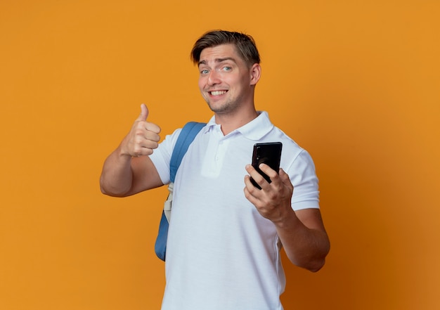 Souriant jeune étudiant beau mâle portant un sac à dos tenant le téléphone et son pouce vers le haut isolé sur orange