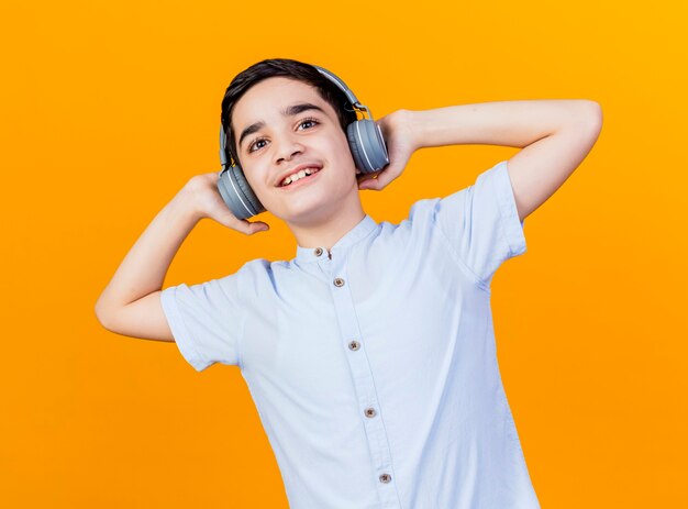Souriant jeune garçon caucasien portant des écouteurs en les touchant en regardant la caméra isolée sur fond orange