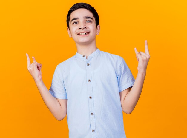 Souriant jeune garçon caucasien regardant la caméra faisant signe de roche isolé sur fond orange