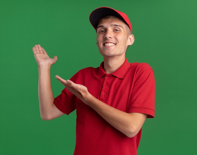 Souriant jeune garçon de livraison blonde points à la main vide isolé sur un mur vert avec espace copie