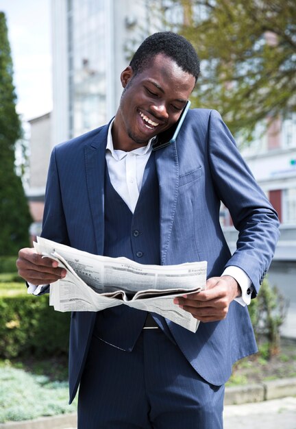 Souriant jeune homme d&#39;affaires parlant sur téléphone portable en lisant le journal