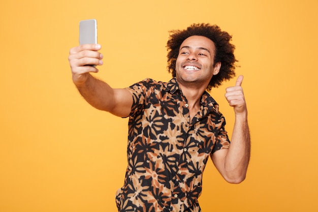 Souriant jeune homme africain faire selfie tout en montrant les pouces vers le haut