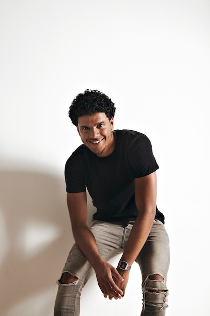 Photo gratuite souriant jeune homme afro-américain athlétique en t-shirt en coton noir blanc et jeans assis à un mur blanc