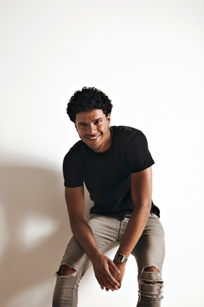 Souriant jeune homme afro-américain athlétique en t-shirt en coton noir blanc et jeans assis à un mur blanc